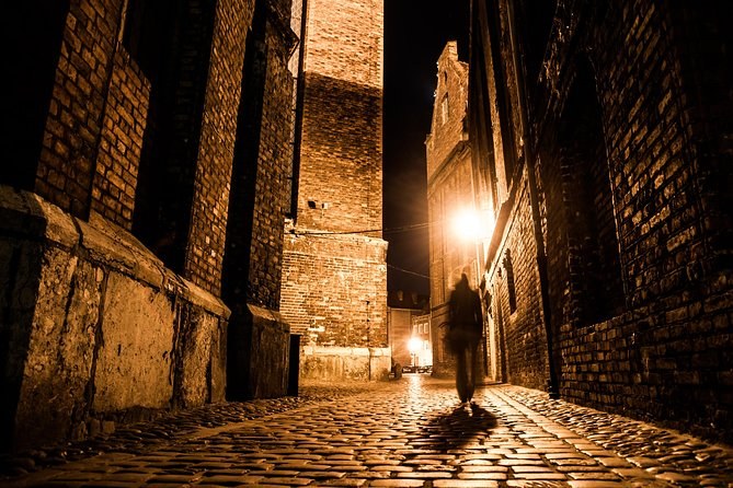A cobbled passage between old brick walls at night, a single figure silhouetted under the one working lamp