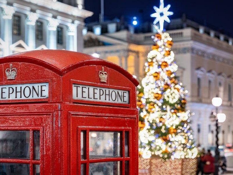 A red London telephone box beside a tall Christmas tree strung with warm white lights after dark