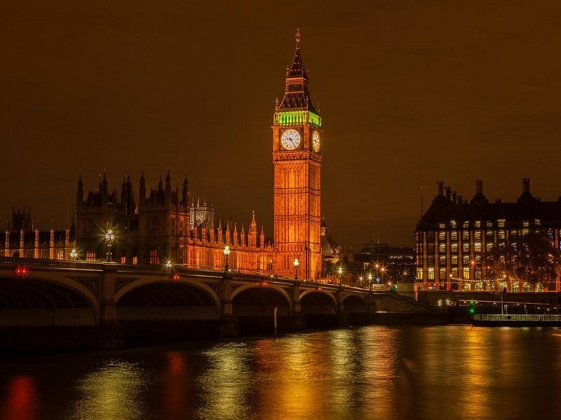 The full length of the Palace of Westminster lit gold at night, seen across the black water of the Thames